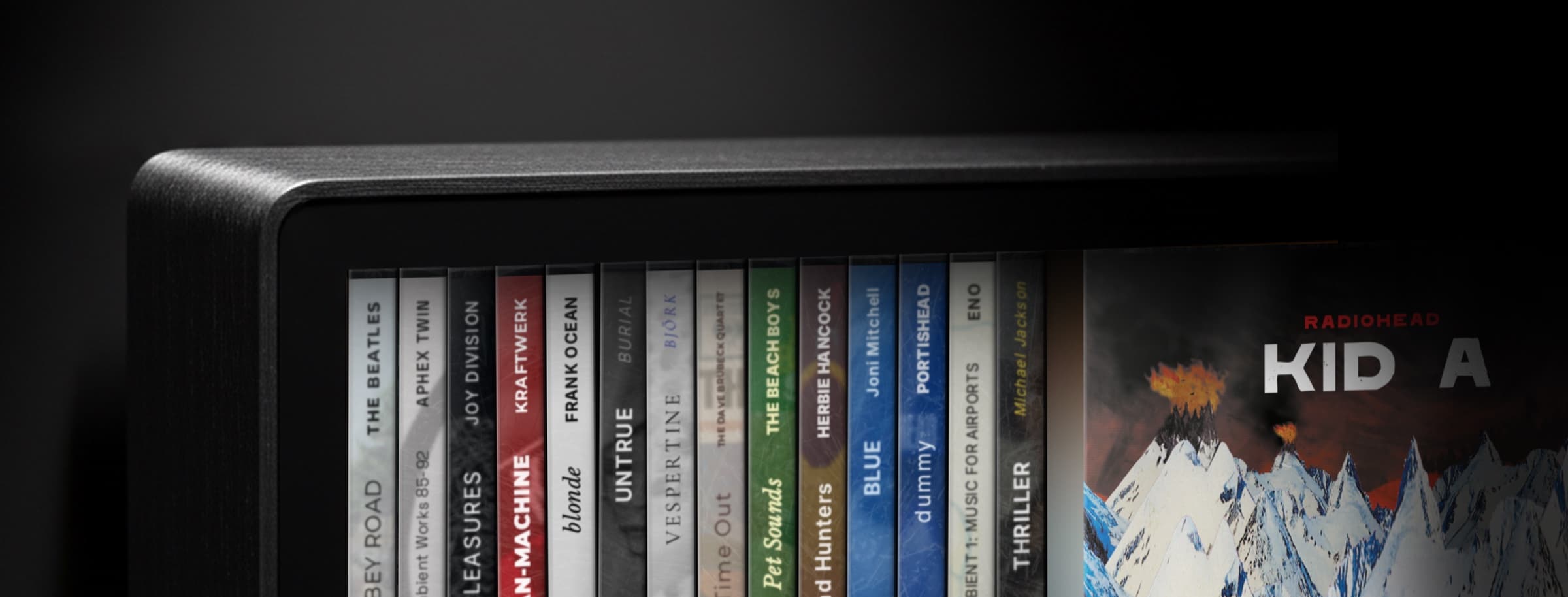 Close-up of the Spine display — brushed aluminum end cap meeting the matte black border, with album spines and Radiohead's Kid A cover visible