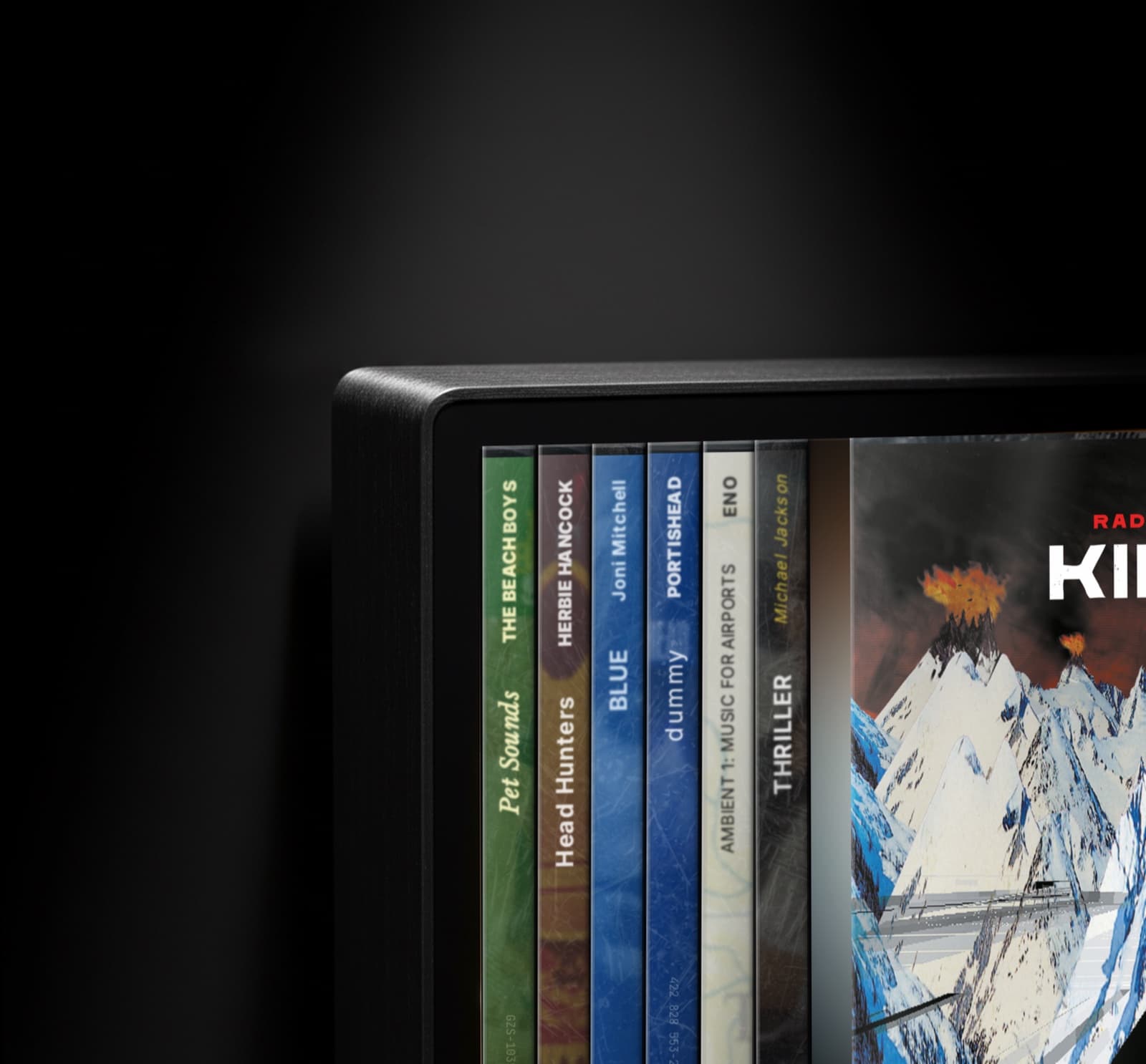 Spine product detail: the rounded brushed-aluminium corner of the device, with album spines visible (Pet Sounds, Head Hunters, Blue, dummy, Portishead, Music for Airports, Thriller) and a Radiohead Kid A album cover on the right.