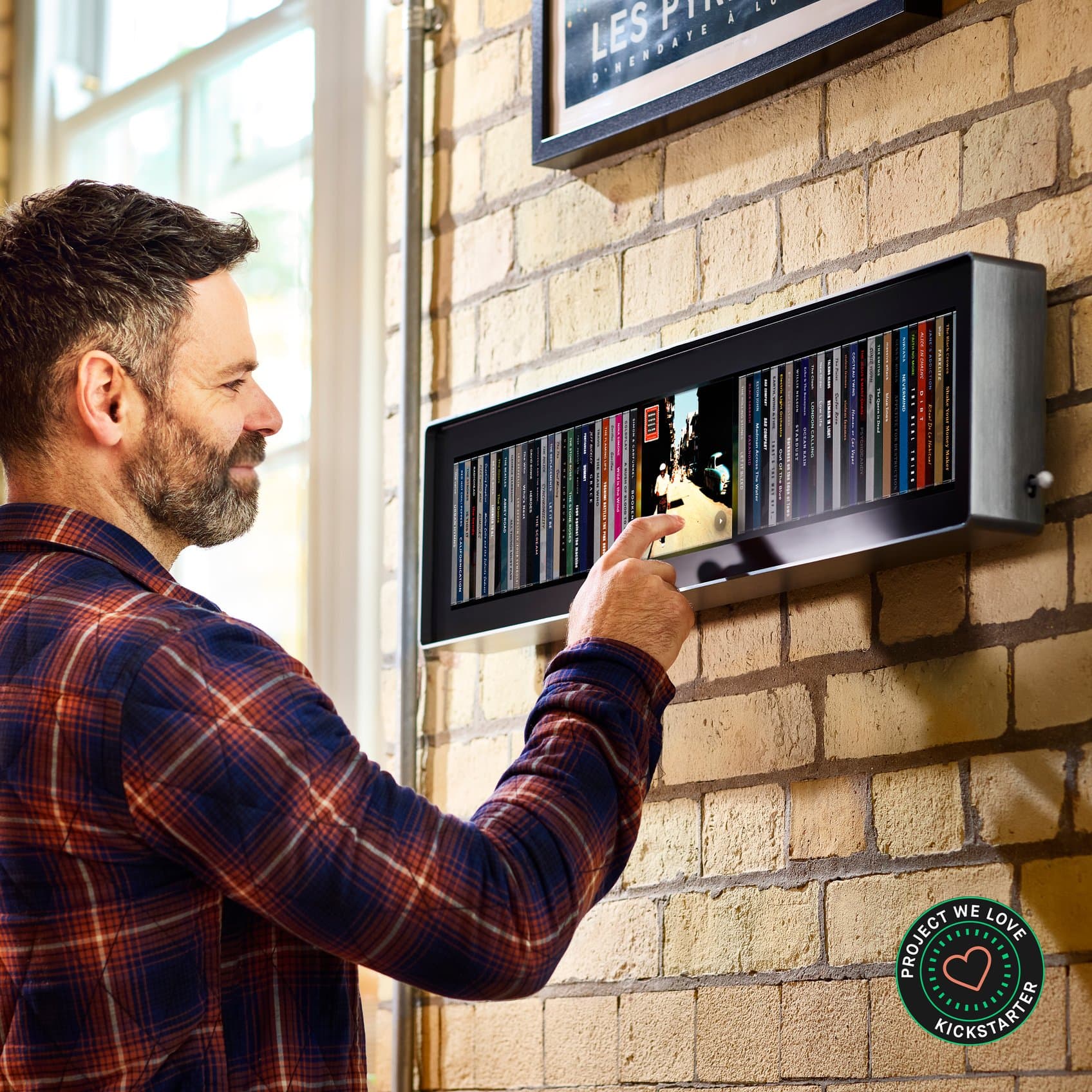 A man touching an album on Spine, mounted on a pale brick wall beside a tall window, with a row of album spines lit across the display. A Kickstarter Project We Love badge sits in the corner.