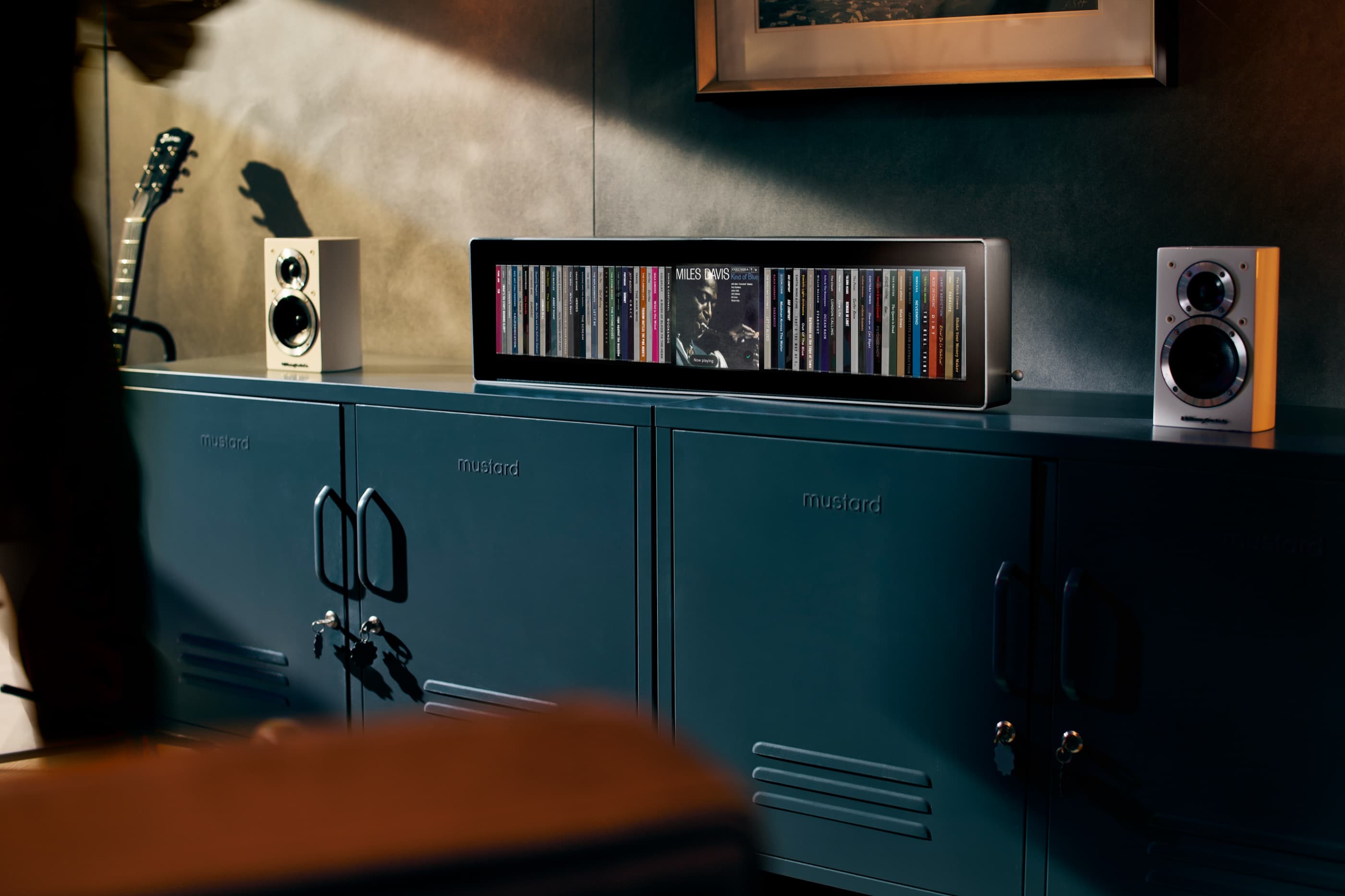 Spine on a dark green metal sideboard between two bookshelf speakers, with an electric guitar to the left and a row of album spines glowing in a softly lit room.