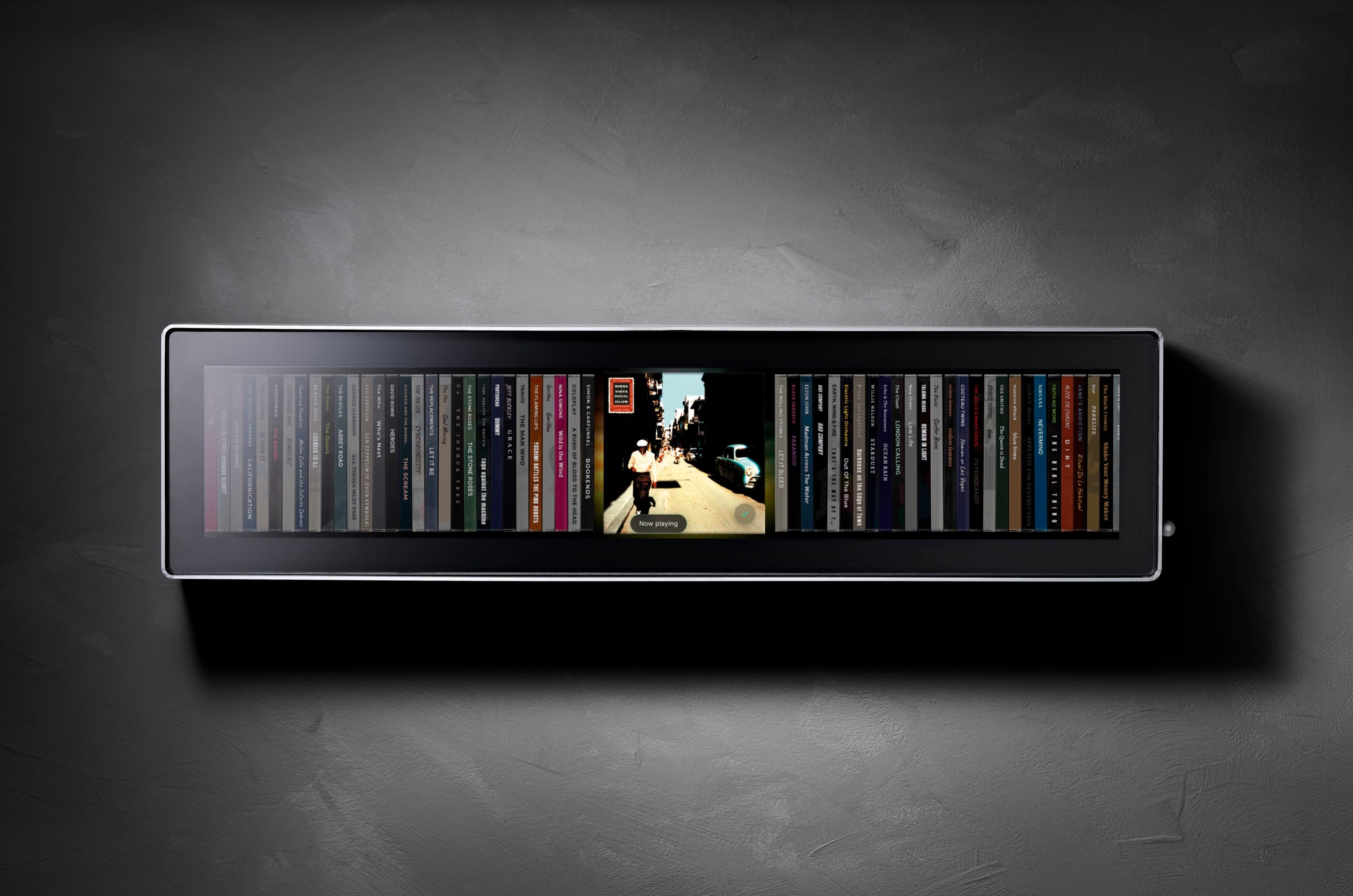 Spine seen front-on, mounted on a dark plaster wall: a brushed aluminium frame around a matte black display showing a row of album spines, with one album cover open in the centre and marked Now playing.