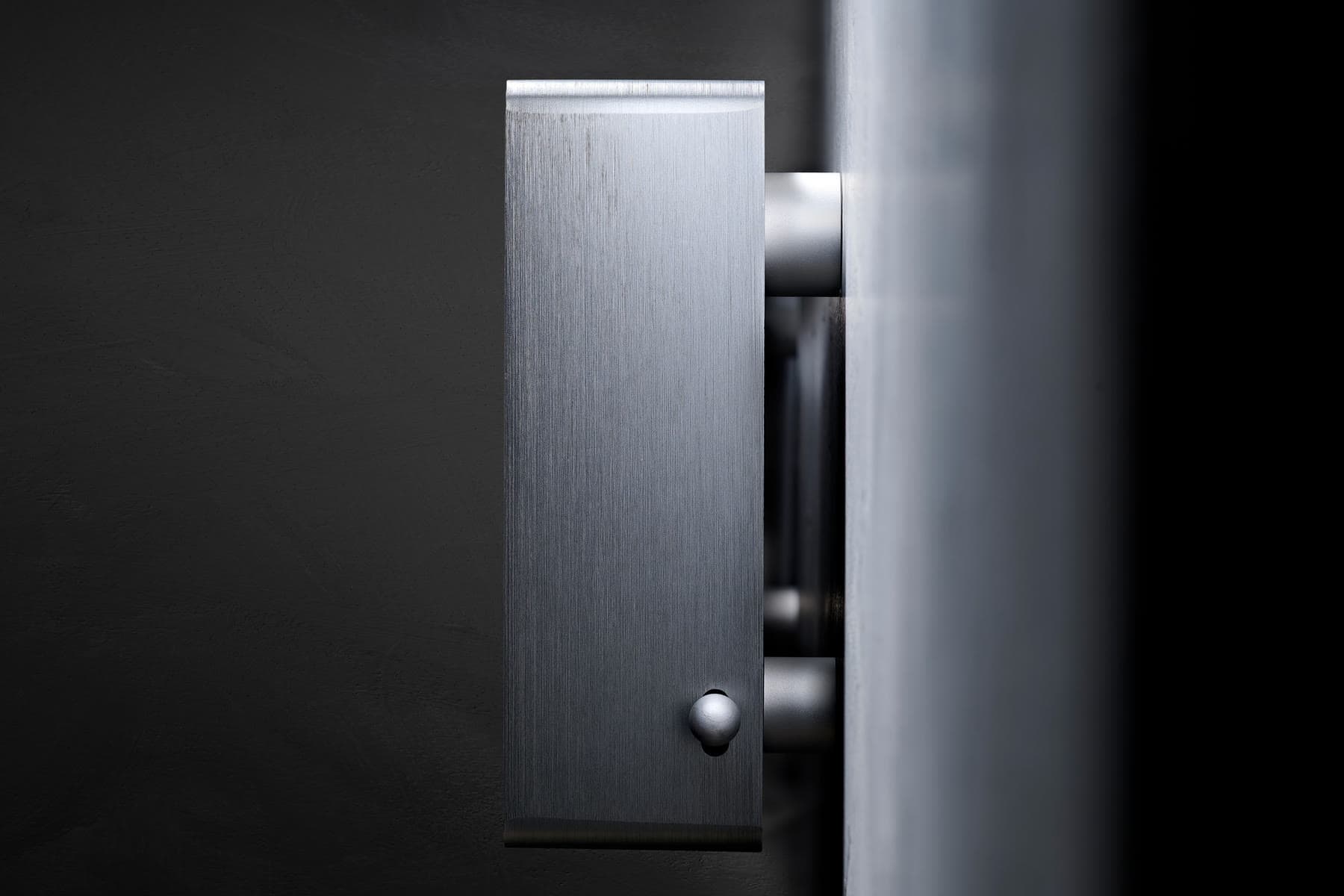 Spine seen side-on against a dark wall: the brushed aluminium end of the body with its small round side switch, held just off the wall on its mount.