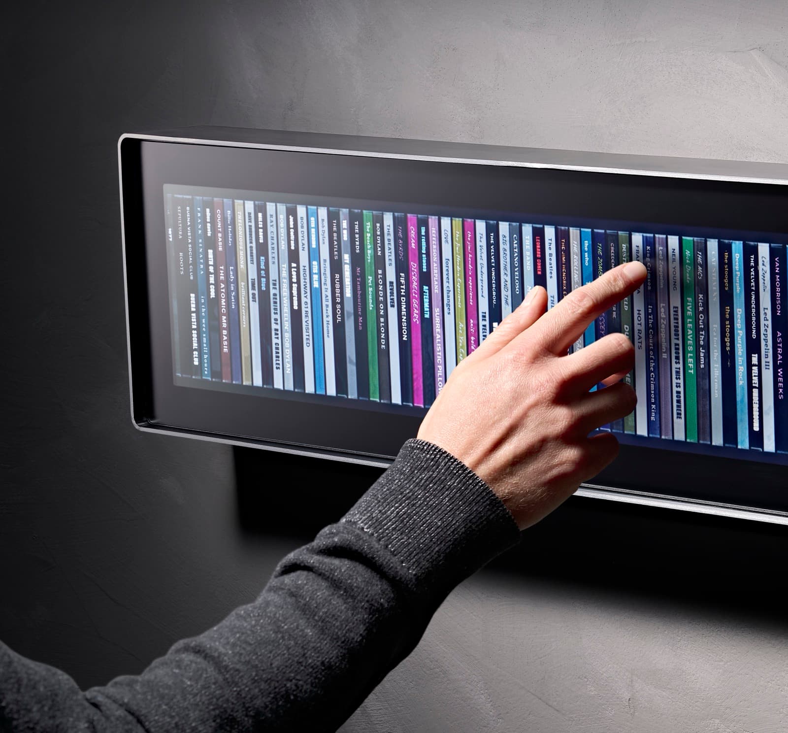 A hand reaching in to touch an album spine on the Spine display, mounted on a dark plaster wall. The left edge of the brushed aluminium body is in view, with a row of album spines lit behind matte glass.