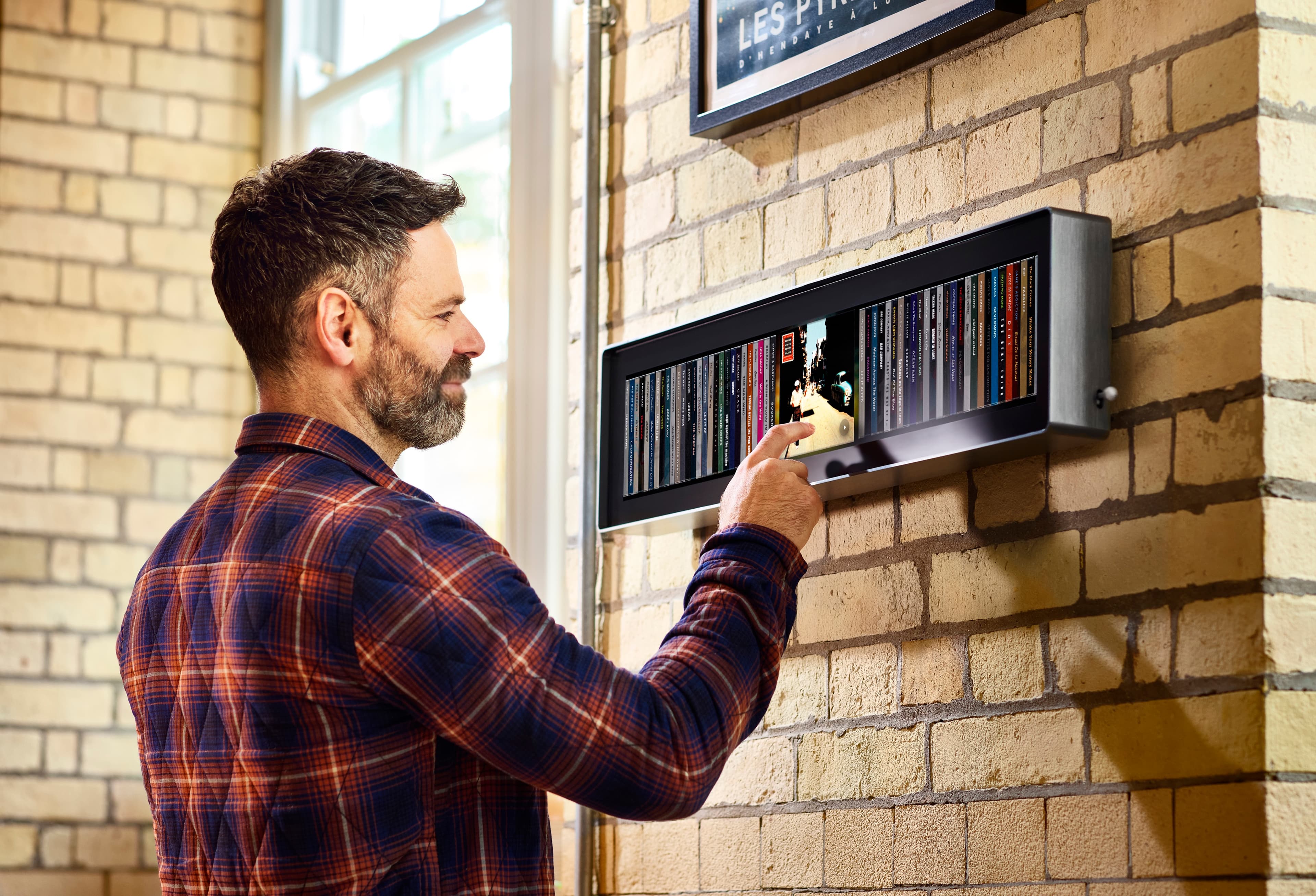 A man touching an album on Spine, mounted on a pale brick wall beside a tall window, with a row of album spines lit across the display