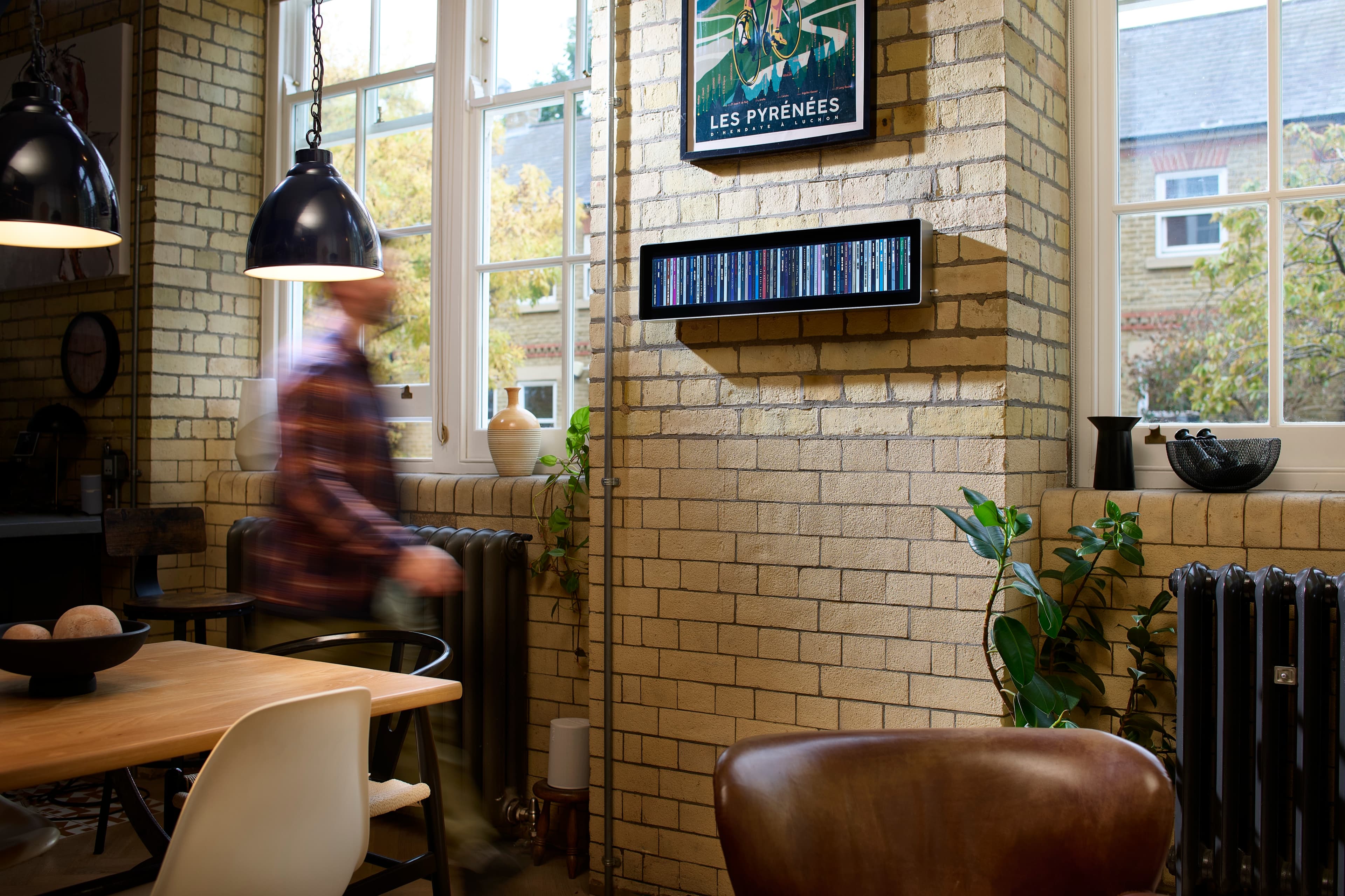 A loft living room with Spine on a pale brick chimney breast beneath a framed poster, a figure walking past in motion blur, pendant lamps and a dining table in the foreground