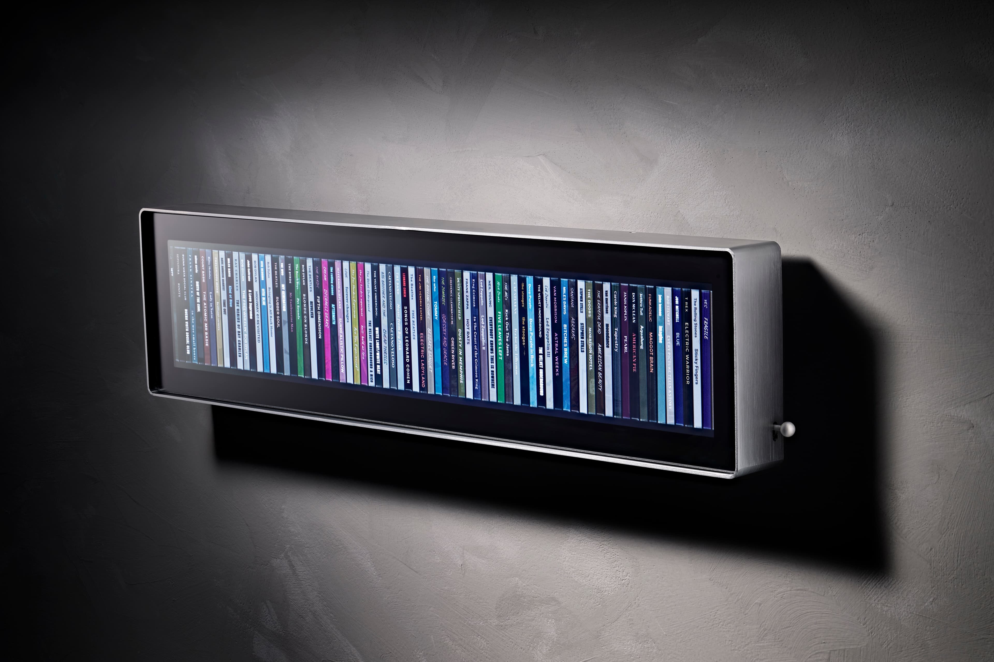 Spine seen from the left at an angle, showing the depth of the brushed aluminium body and a full row of album spines behind matte glass
