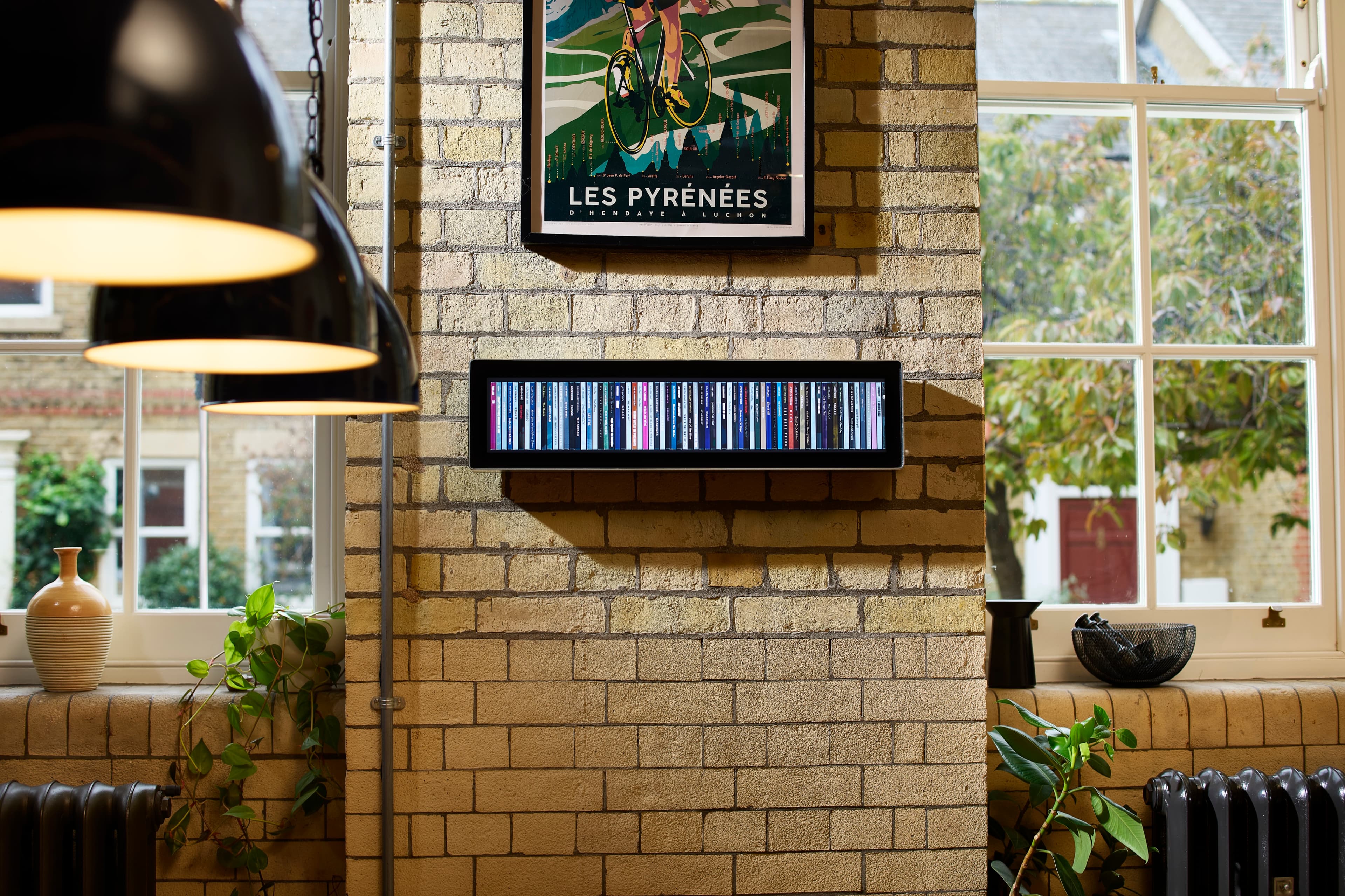 Spine mounted on a pale brick wall between two tall windows, beneath a framed cycling poster, with pendant lamps hanging in the foreground