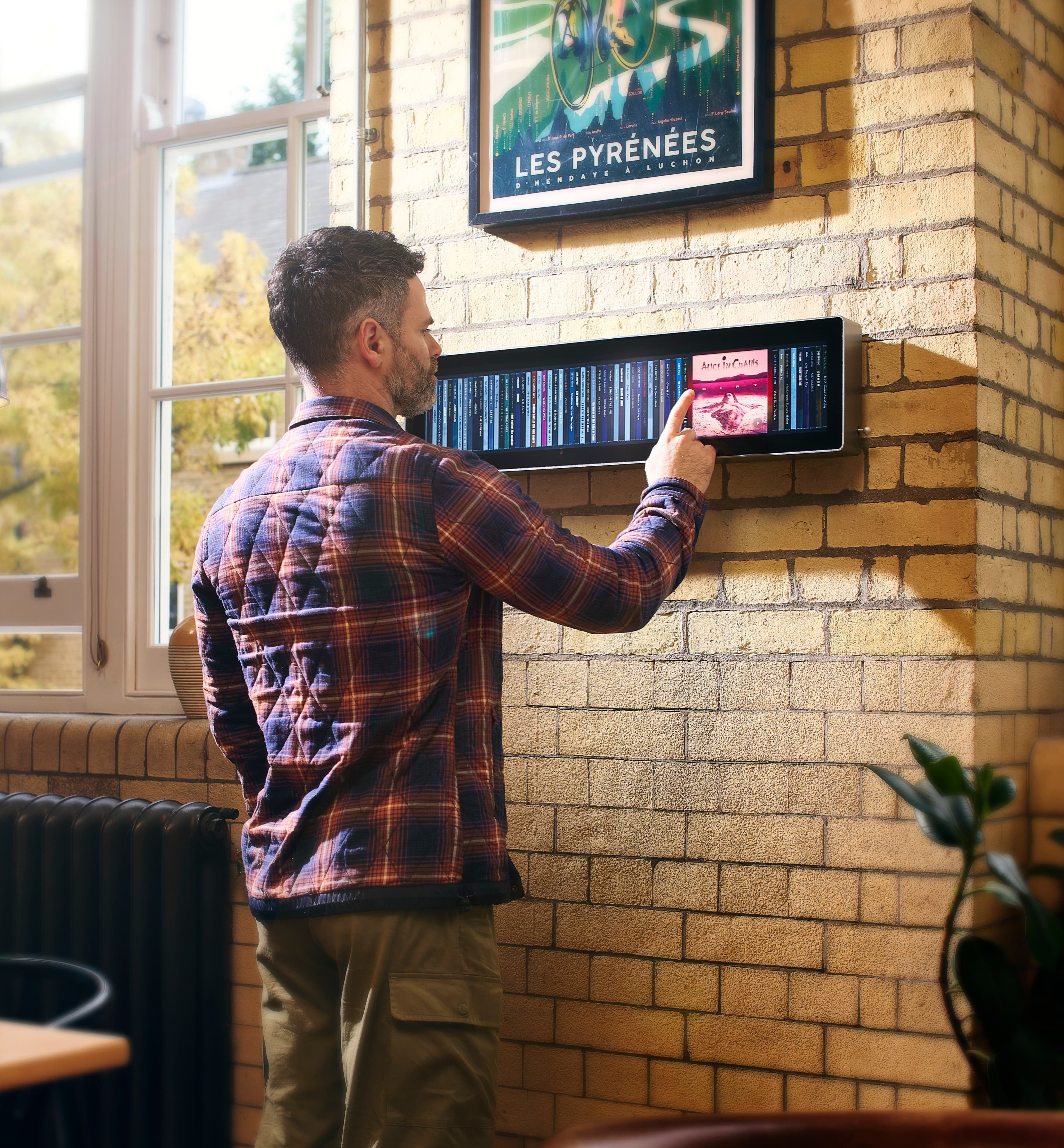 A man in a checked shirt reaching up to tap an album on Spine, mounted on a pale brick wall beside a window