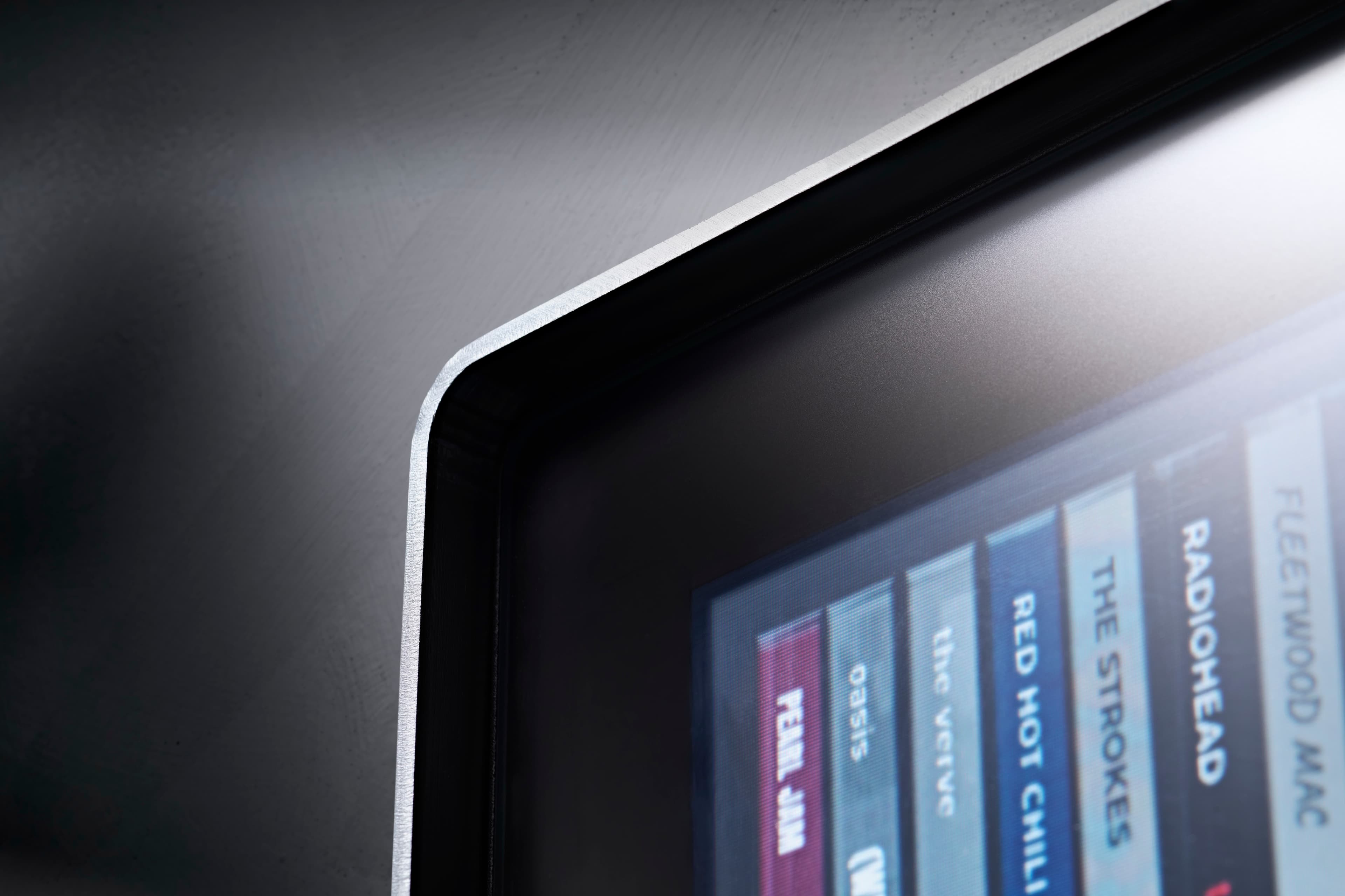 Close-up of Spine's top corner, where the matte glass meets the thin aluminium edge, with album spines in soft focus