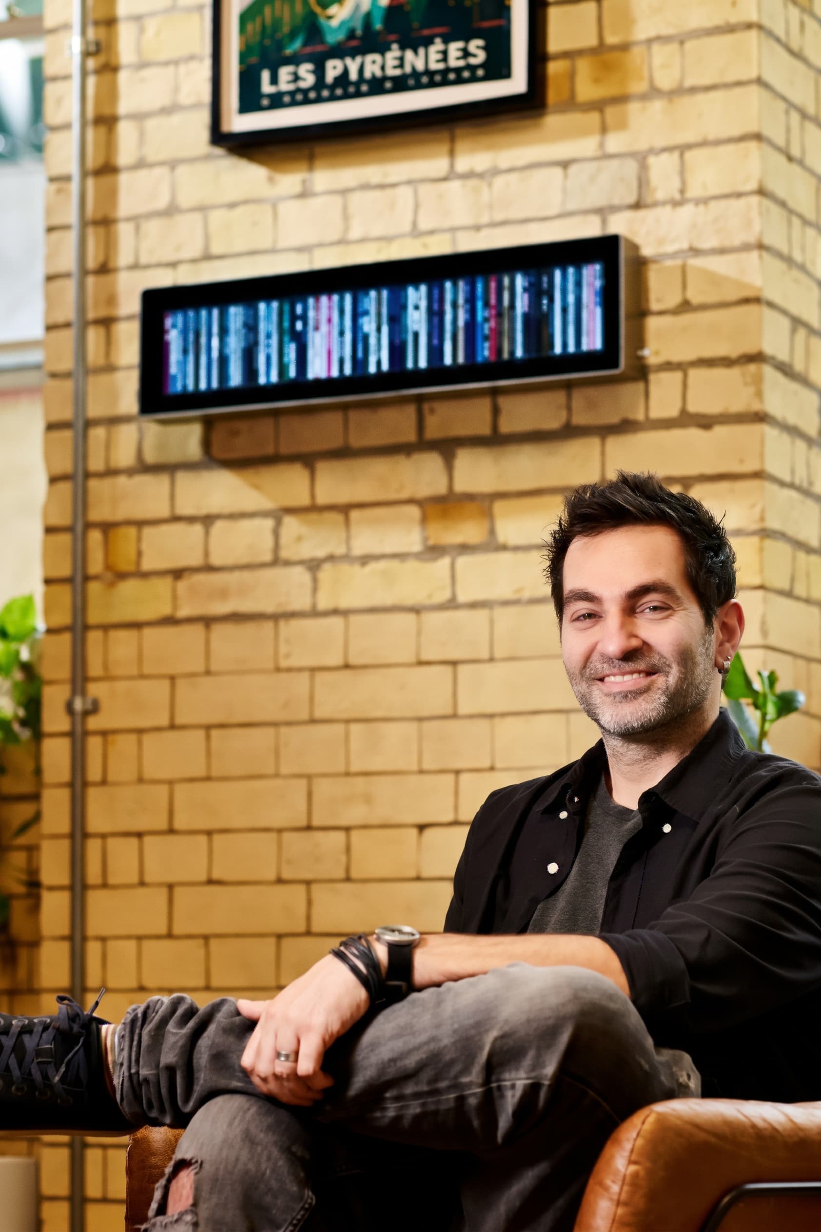 Ricardo Amorim, founder of Spine, smiling in a leather armchair in front of a pale brick wall, with Spine mounted on the wall behind him beneath a framed poster